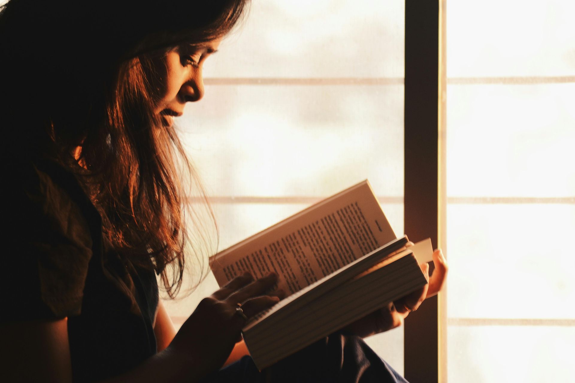 Woman reading a book by a window; sunlight illuminates her face and the pages.