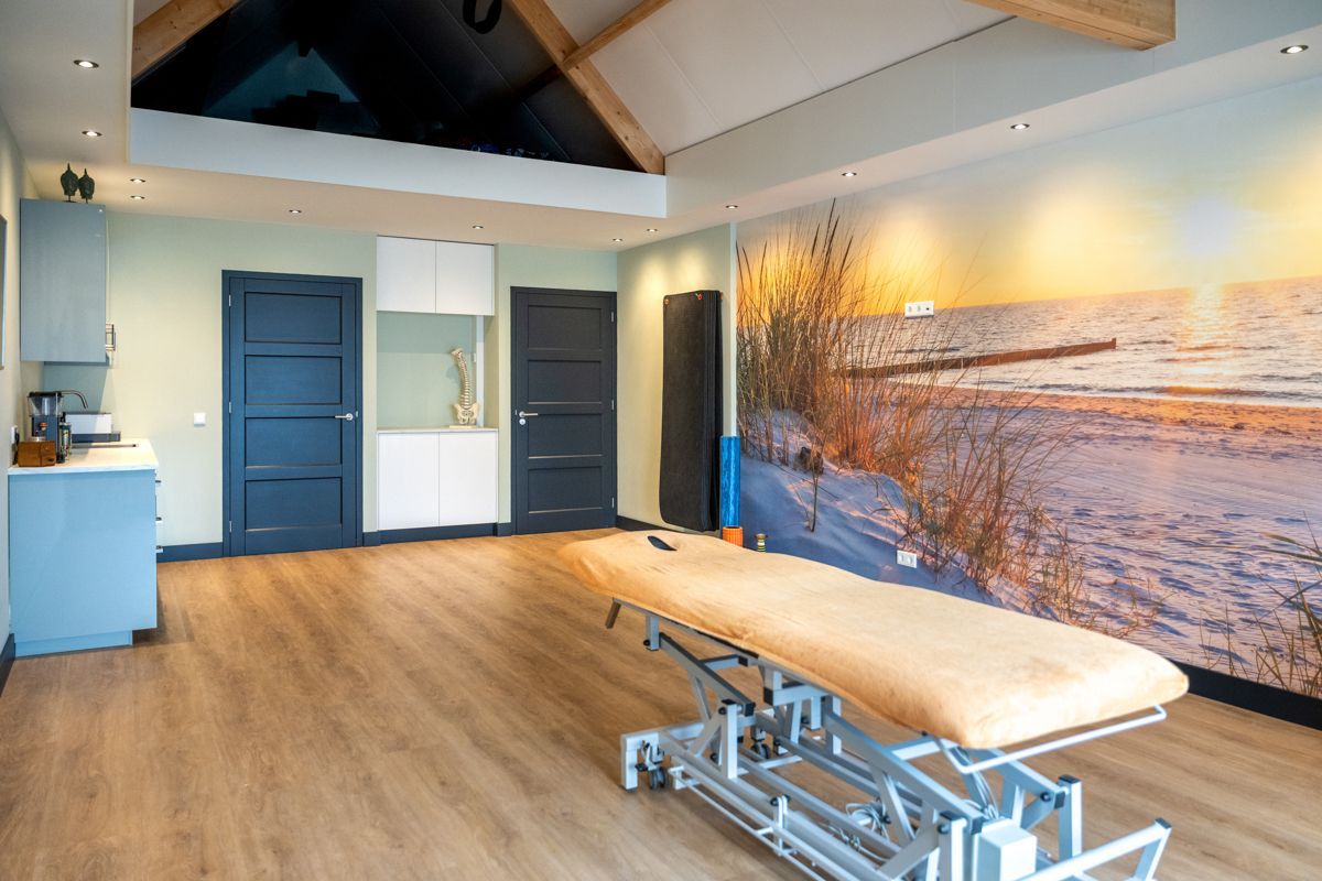 Therapieruimte met behandelbank, strandmuurschildering, blauwe deuren en houten vloer.