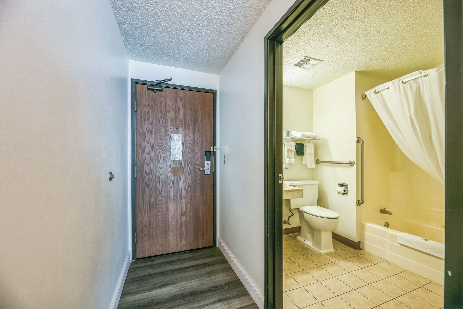 Hallway leading to a hotel room door and bathroom. The bathroom has a toilet, sink, and bathtub.