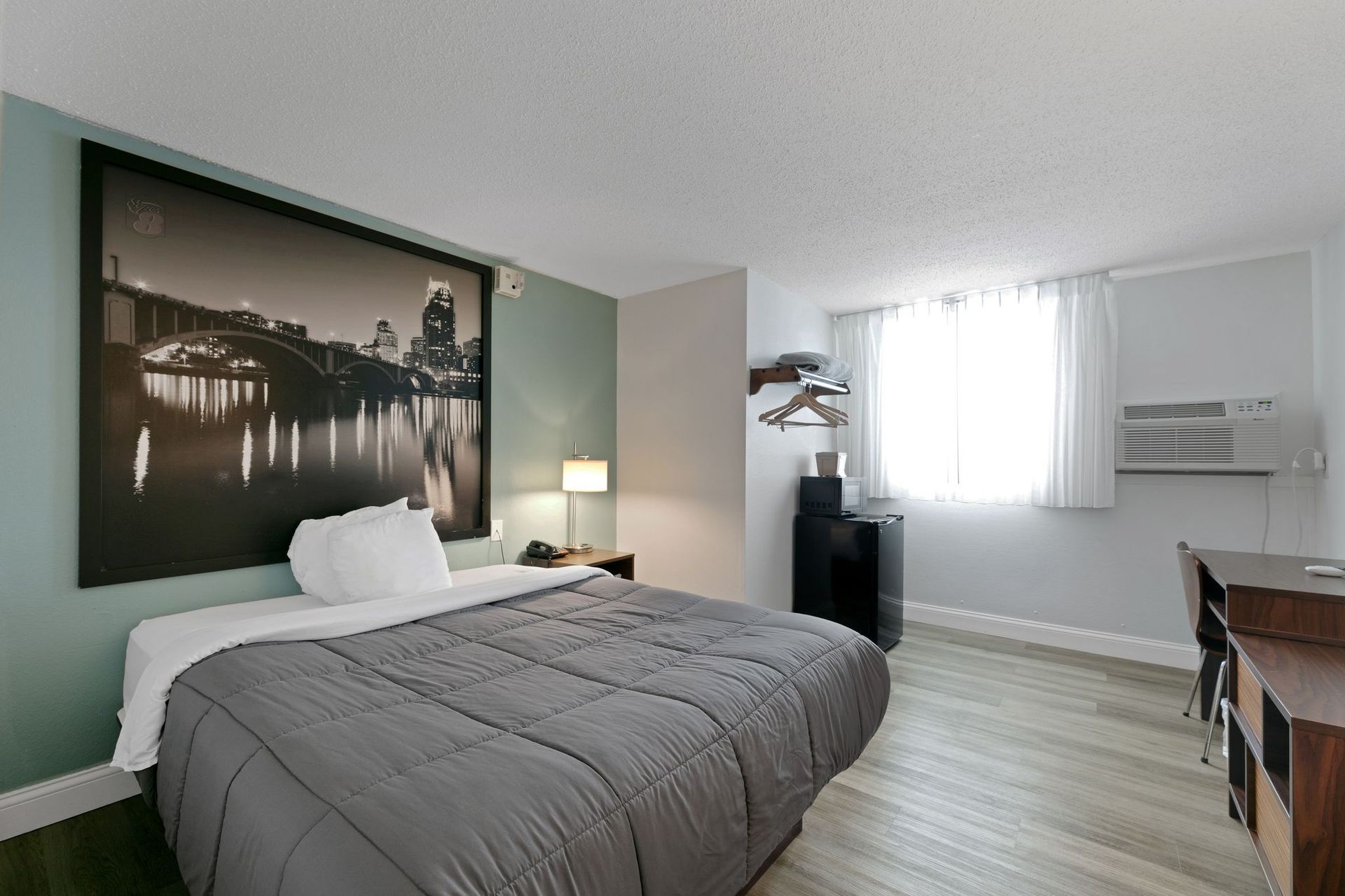 A hotel room with a bed, desk, and city skyline art. Gray bedding, light walls, and a window.