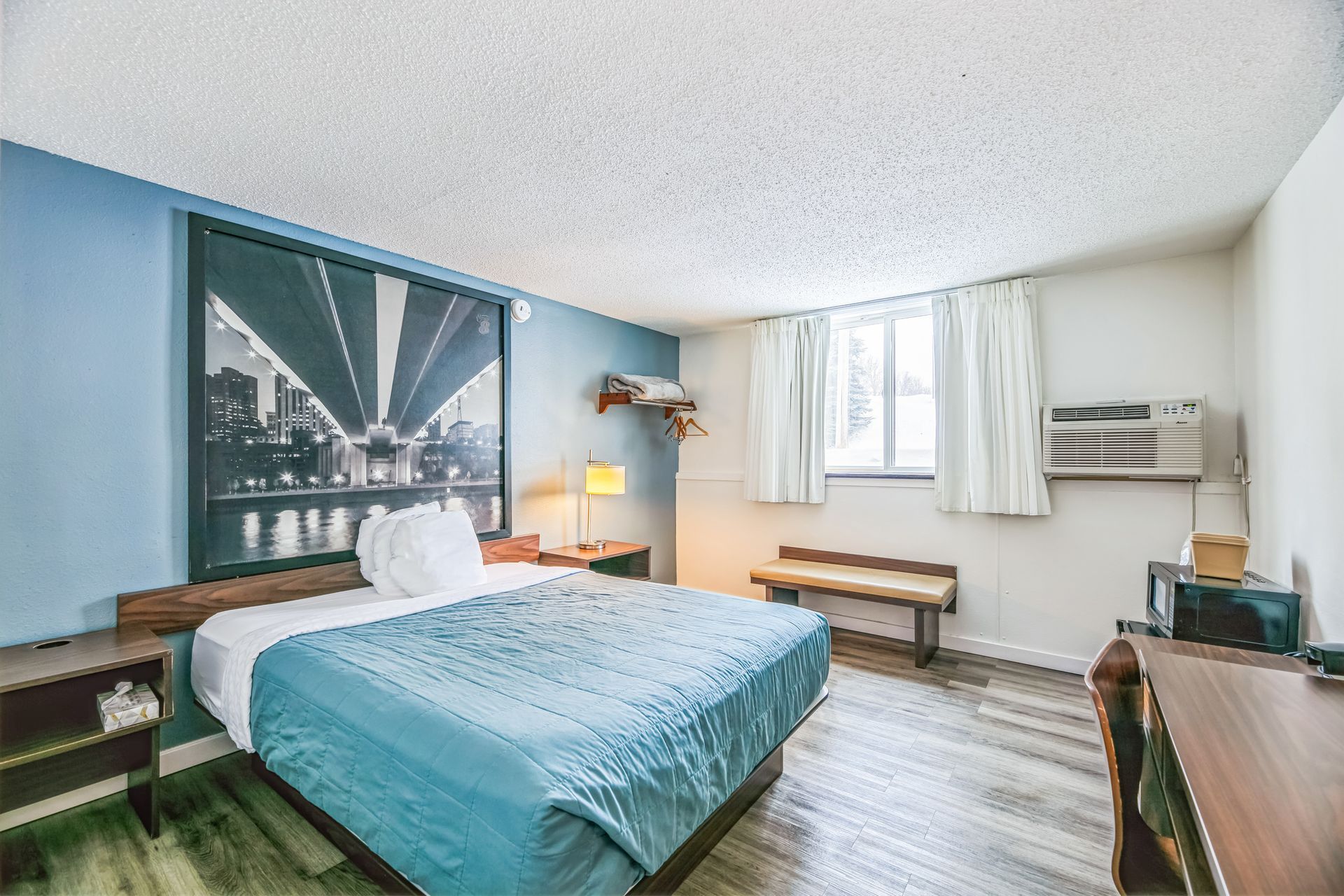 Hotel room with a bed, bench, artwork, window, and microwave. Blue and white walls, wood accents.