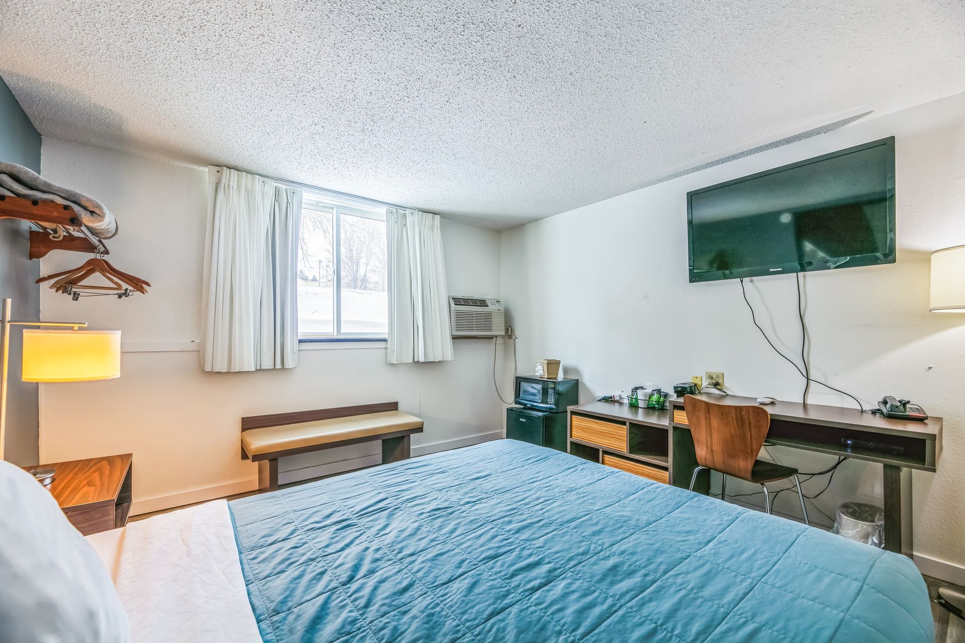 Bedroom with bed, desk, TV, window, and small refrigerator. White walls, blue bedspread.