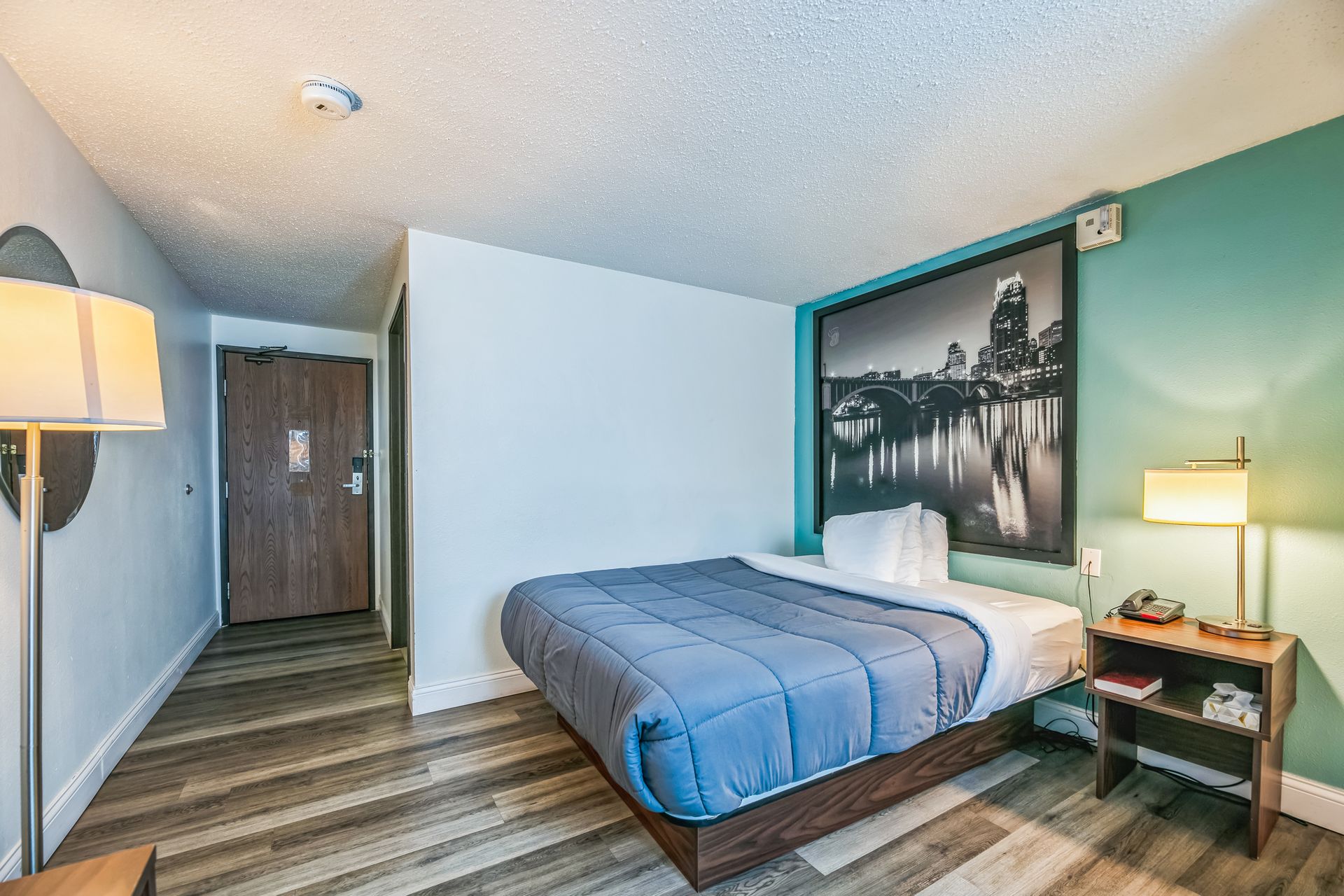 Bedroom with bed, lamp, artwork, and a small nightstand. Includes turquoise wall and wood flooring.