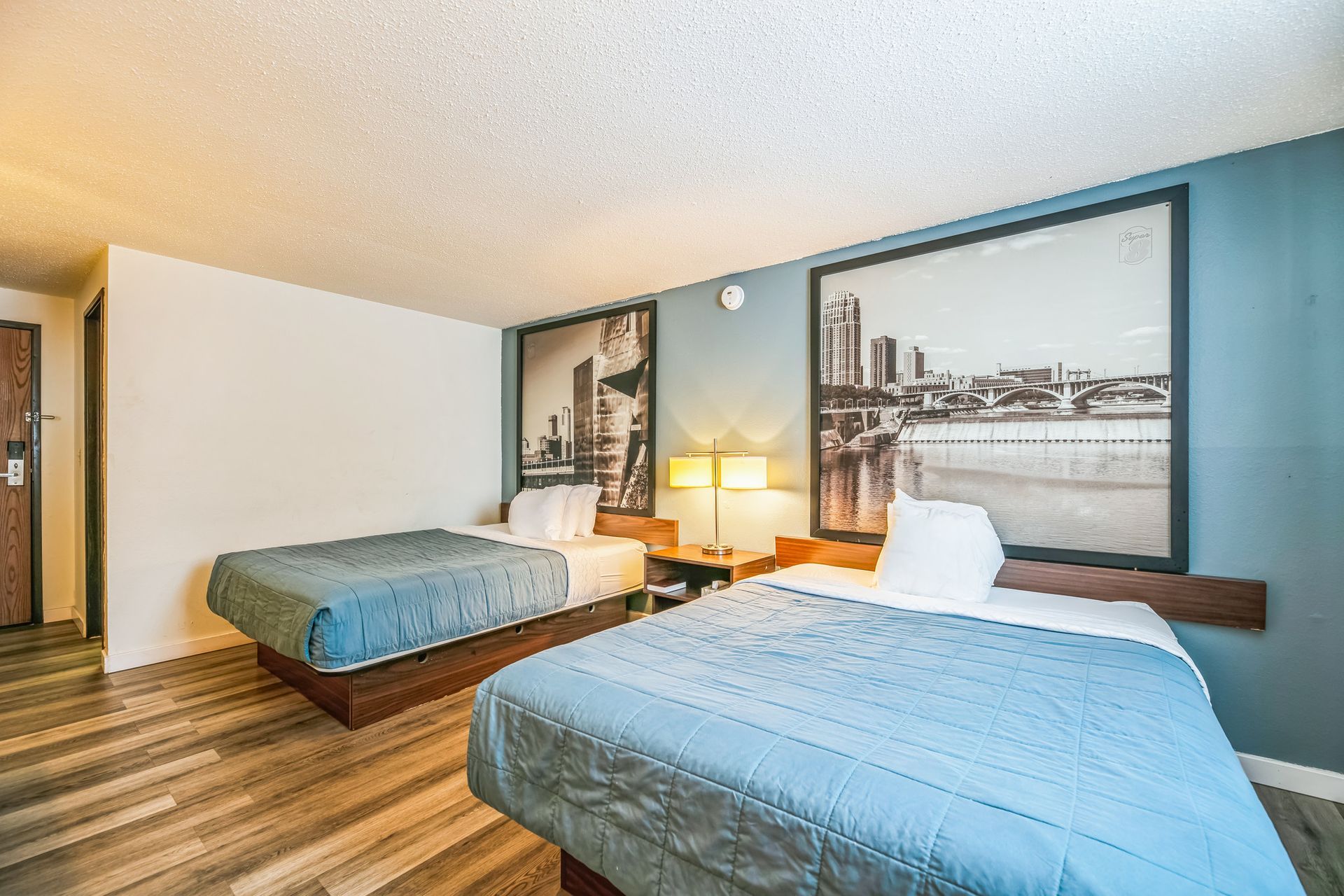 Two beds in a hotel room with light blue comforters. City skyline art hangs above the beds.