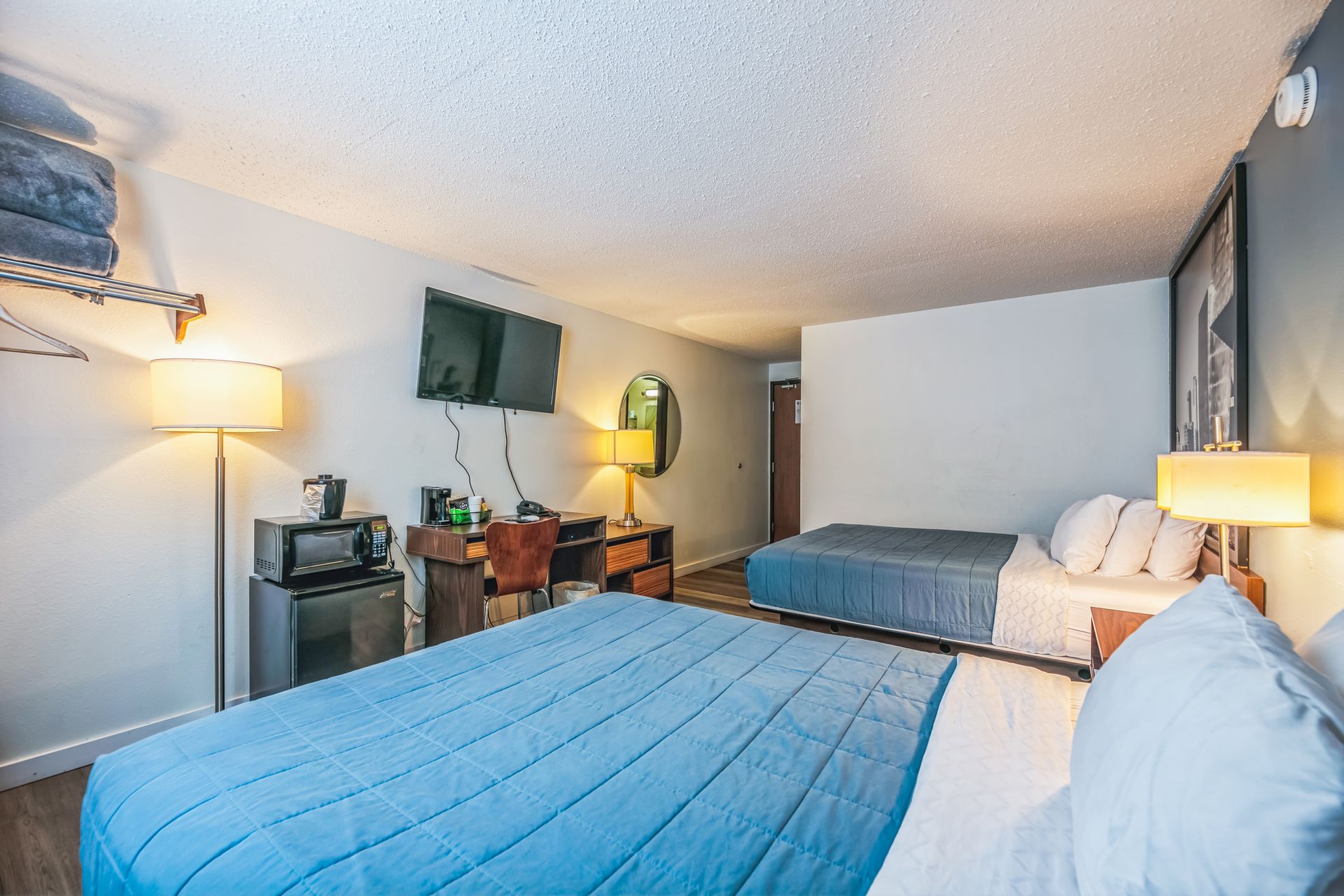 Hotel room with two beds, desk, TV, and mini-fridge. The room has light blue bedding and white walls.
