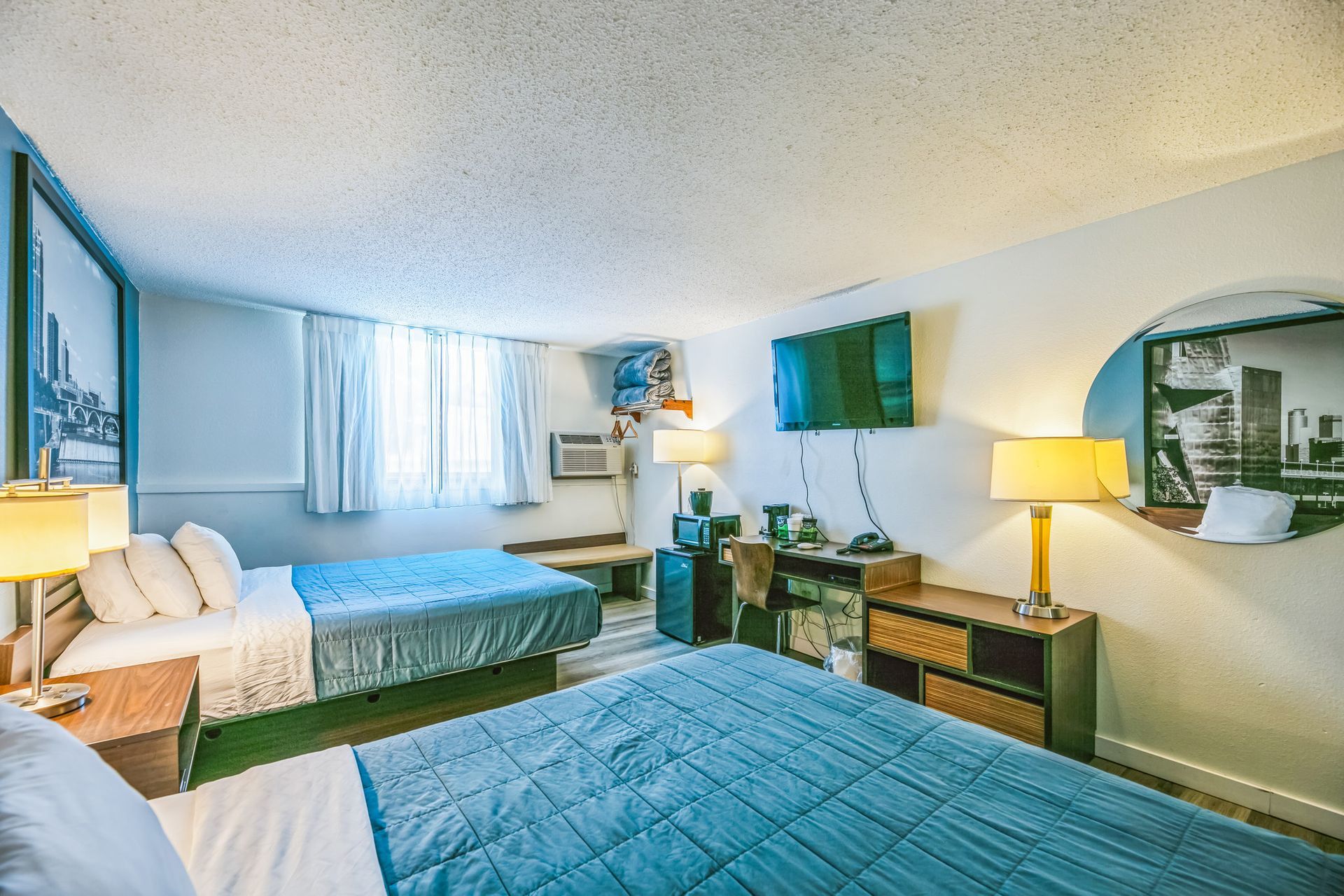 Hotel room with two beds, TV, desk, and round mirror; blue and white color scheme.