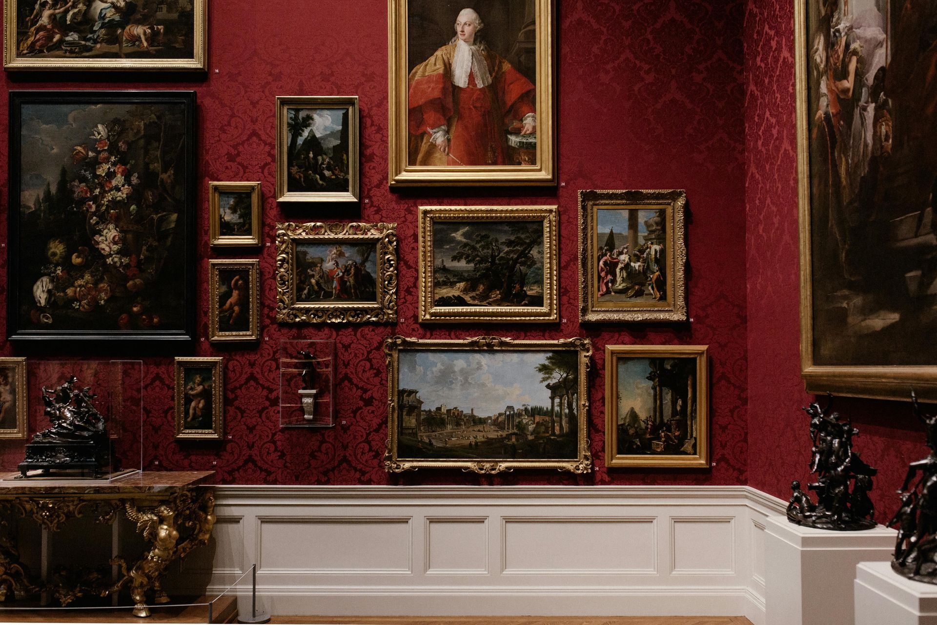 Art gallery with numerous framed paintings on red patterned wallpaper above white wainscoting.