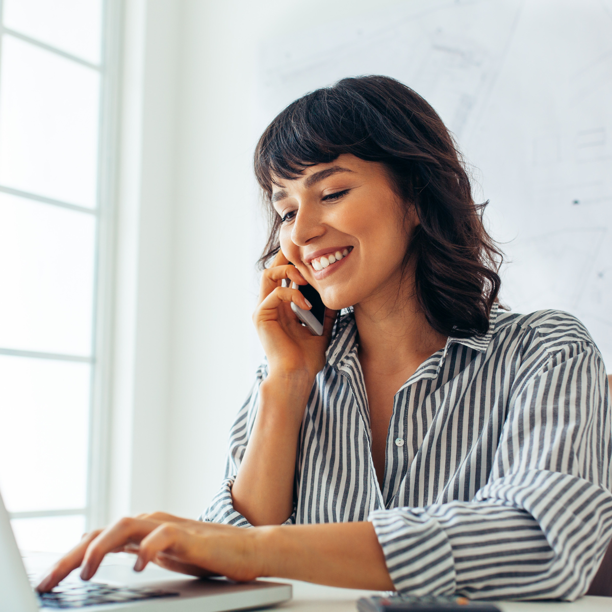 A woman is talking on a cell phone while using a laptop computer.