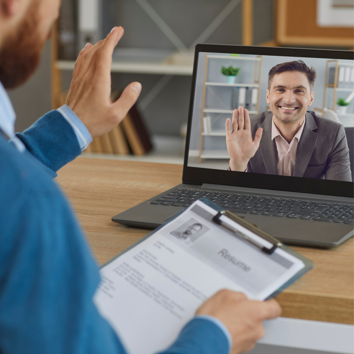 A man is waving at another man on a laptop screen while holding a clipboard.