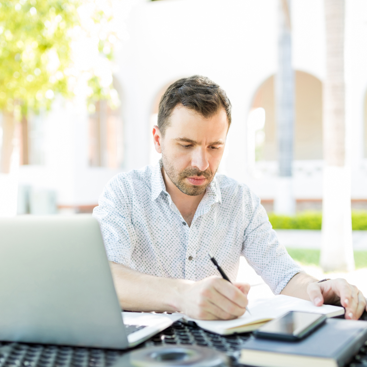 A man is writing in a notebook while using a laptop