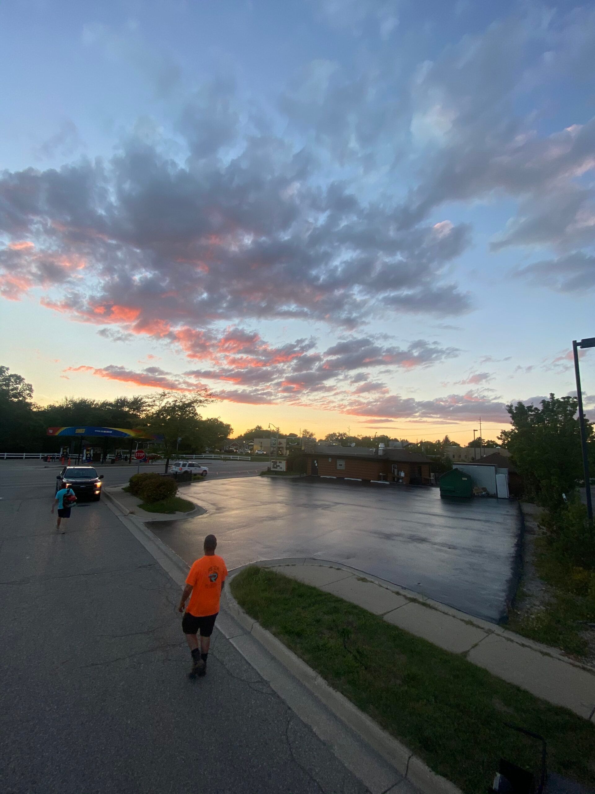 A man in an orange shirt is walking down a street at sunset
