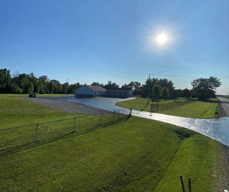 A concrete driveway leading to a house in the middle of a grassy field.