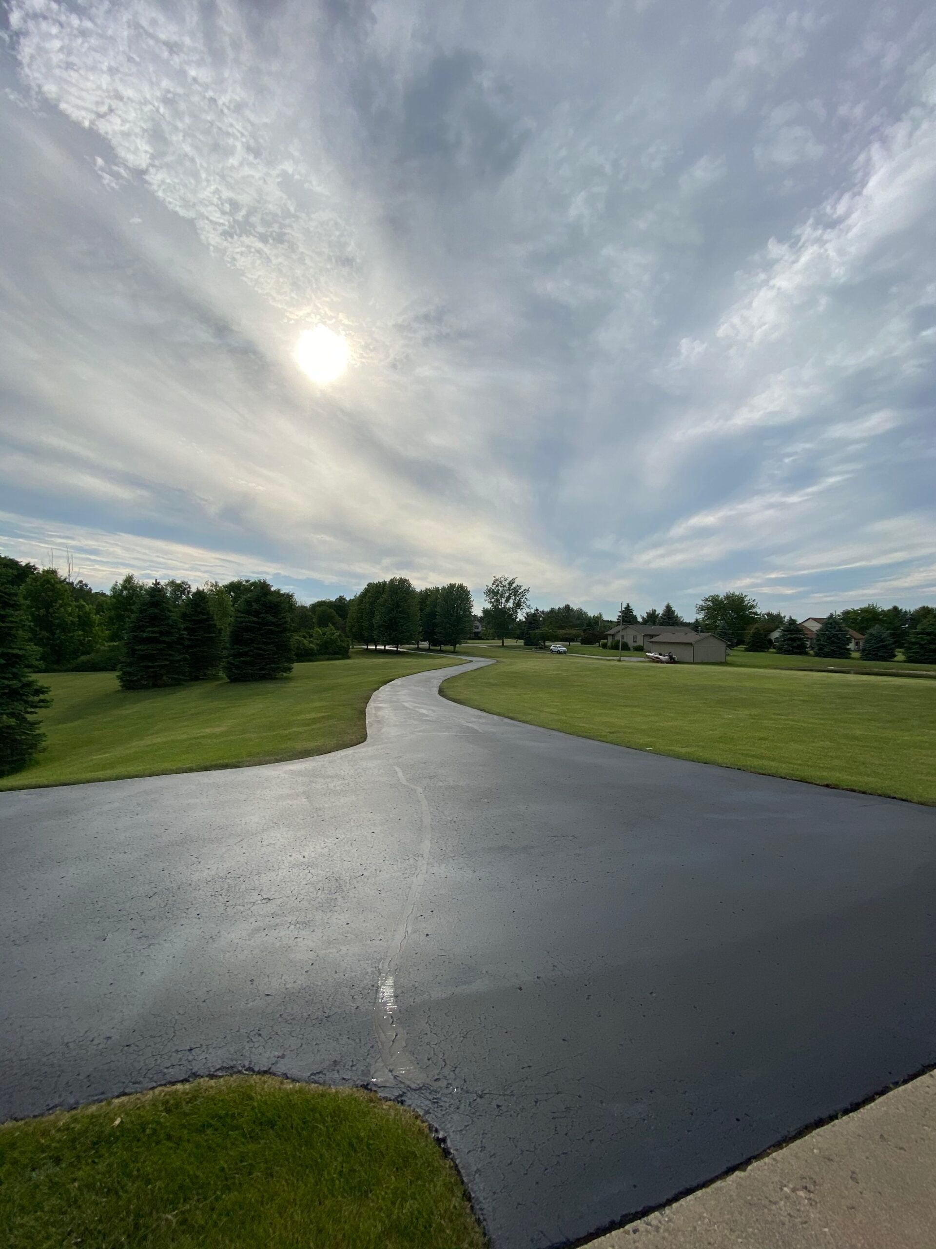 A curvy road going through a grassy field with the sun shining through the clouds.