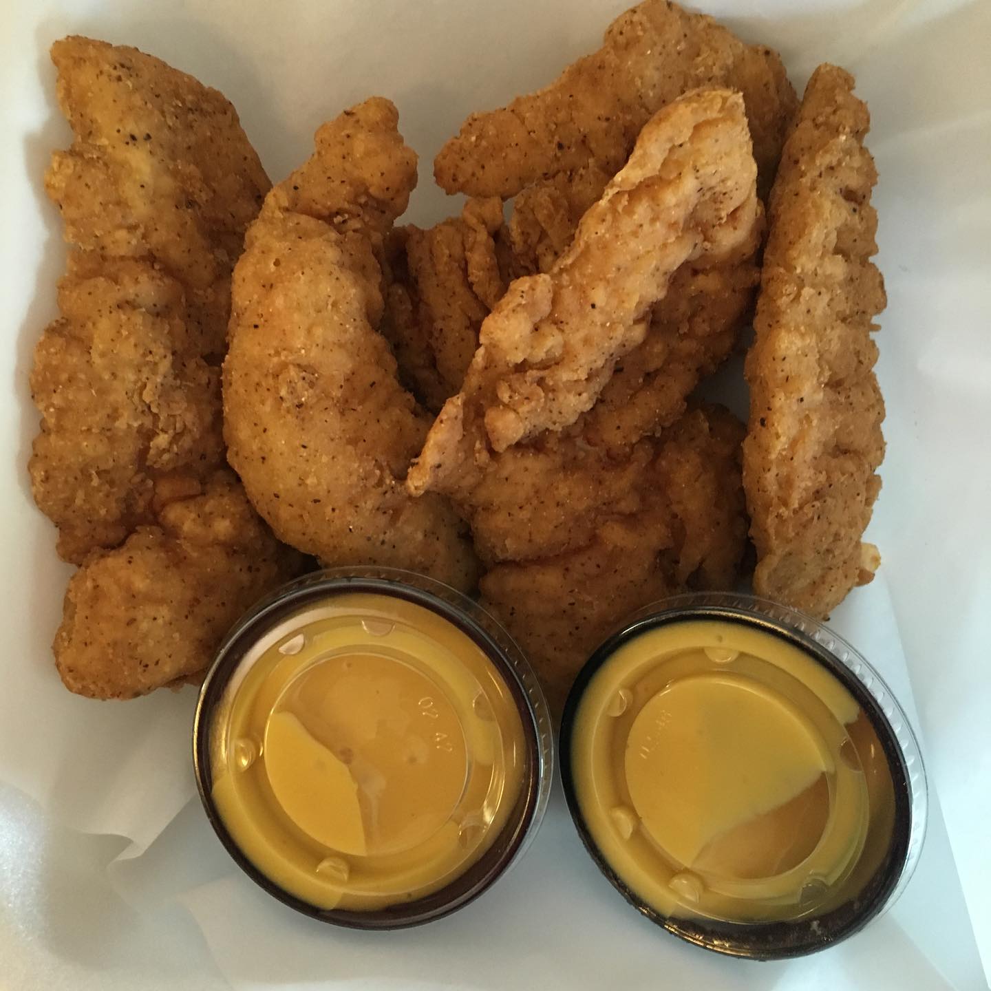 Fried chicken tenders with two containers of yellow dipping sauce in a white container.