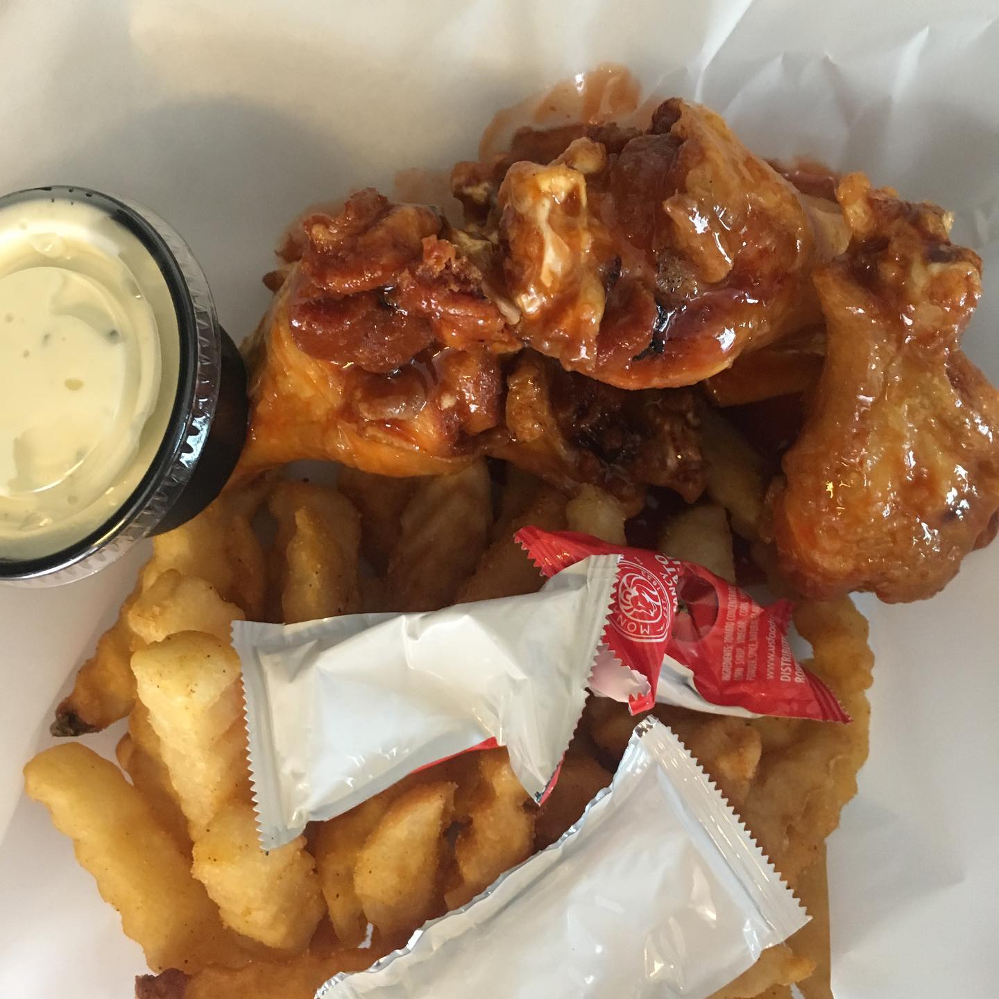 Chicken wings with fries, dipping sauce, and ketchup packets in a white takeout container.