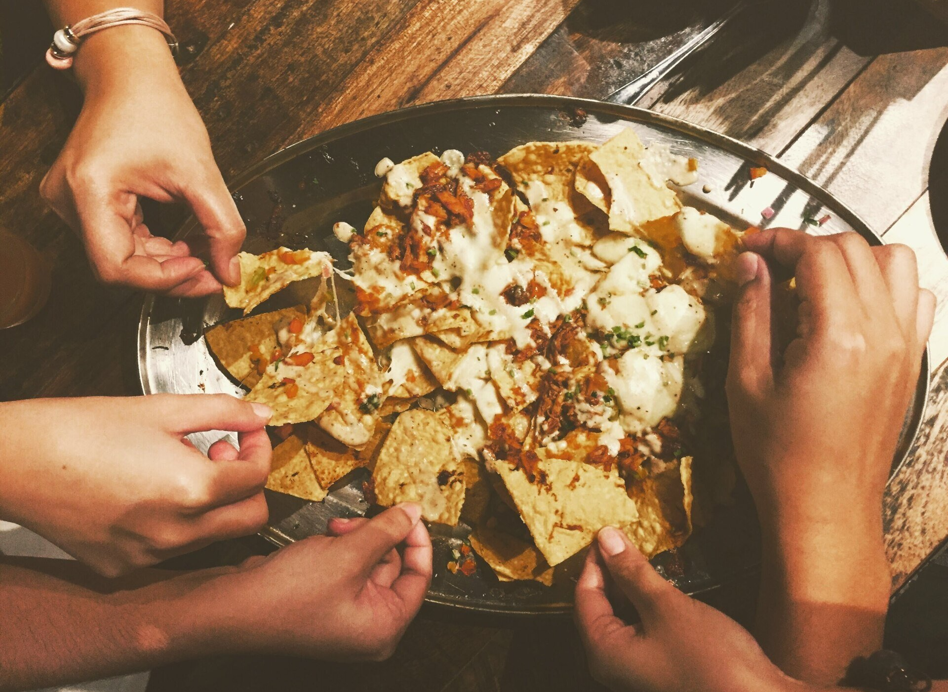 Hands reaching for a plate of loaded nachos on a wooden table.