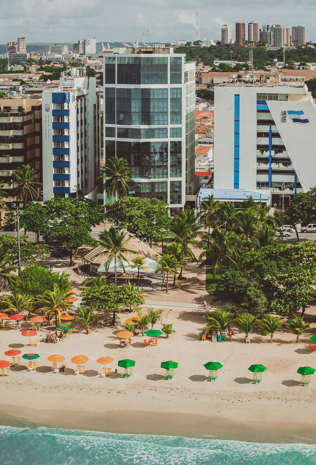 Deck de madeira com mesas, cadeiras e redes sob um guarda-sol perto da praia com palmeiras.