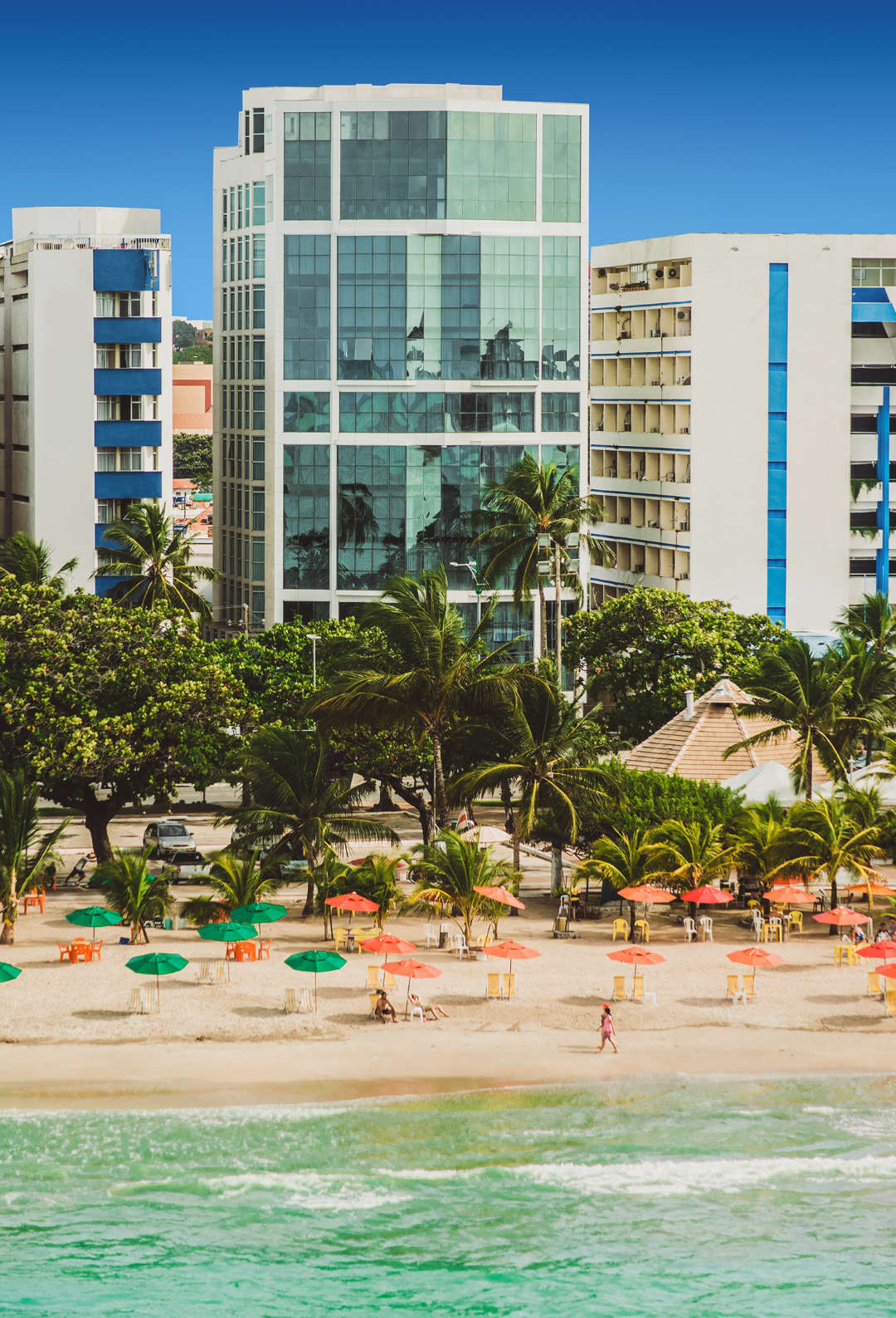 Cena de praia com edifícios, palmeiras e guarda-sóis.