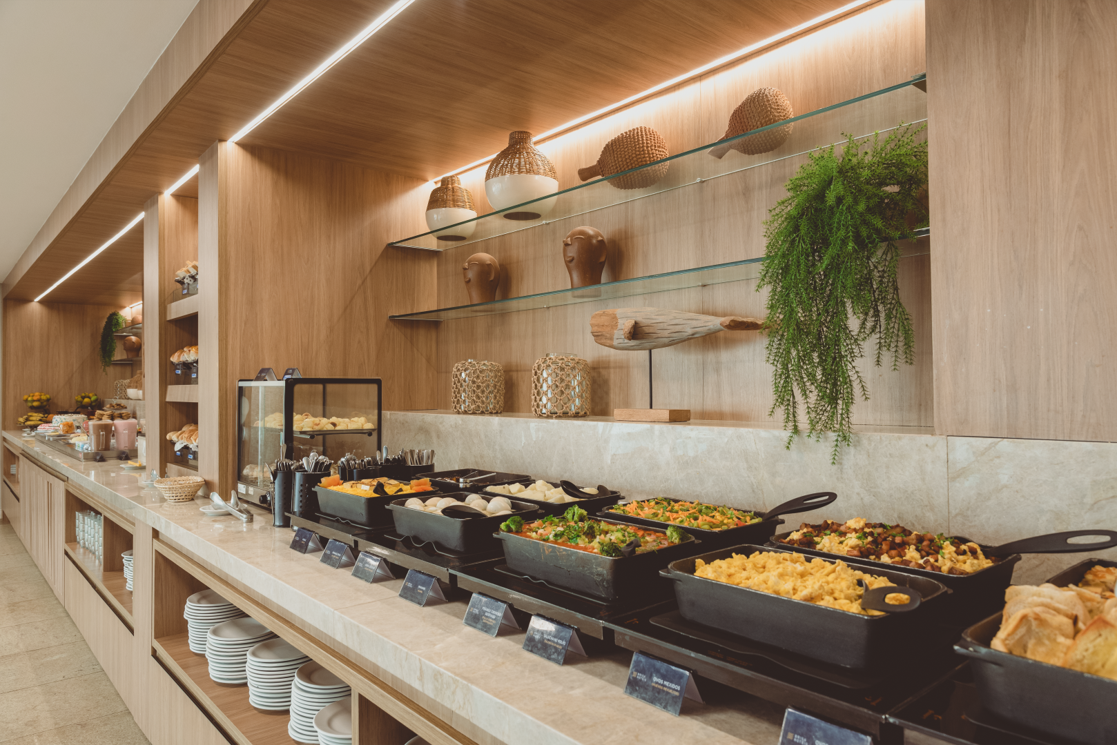 Buffet com diversas opções de comida, prateleiras de madeira com decoração e uma área de jantar moderna e bem iluminada.