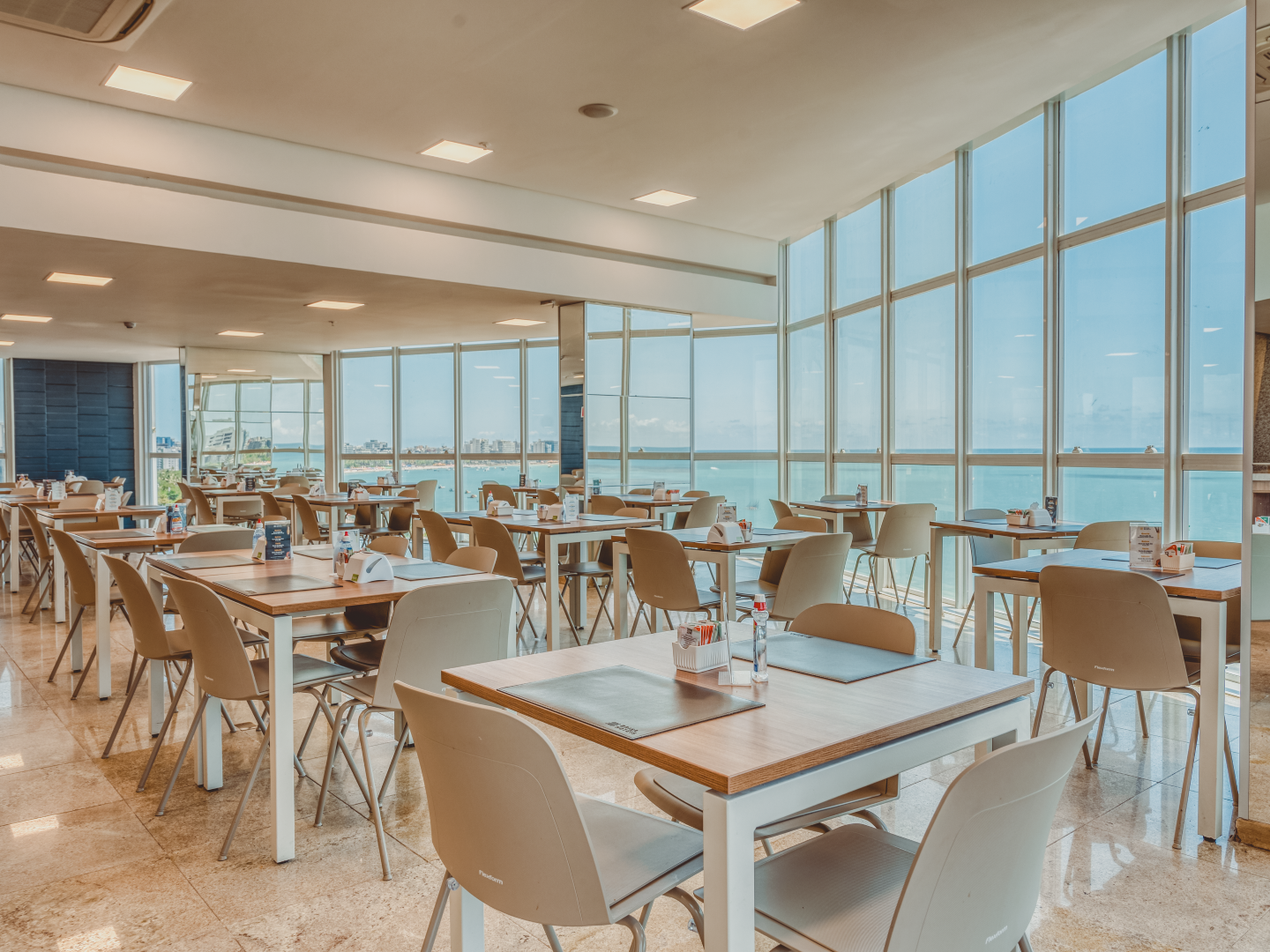 Restaurante com mesas e cadeiras, grandes janelas com vista para o oceano.