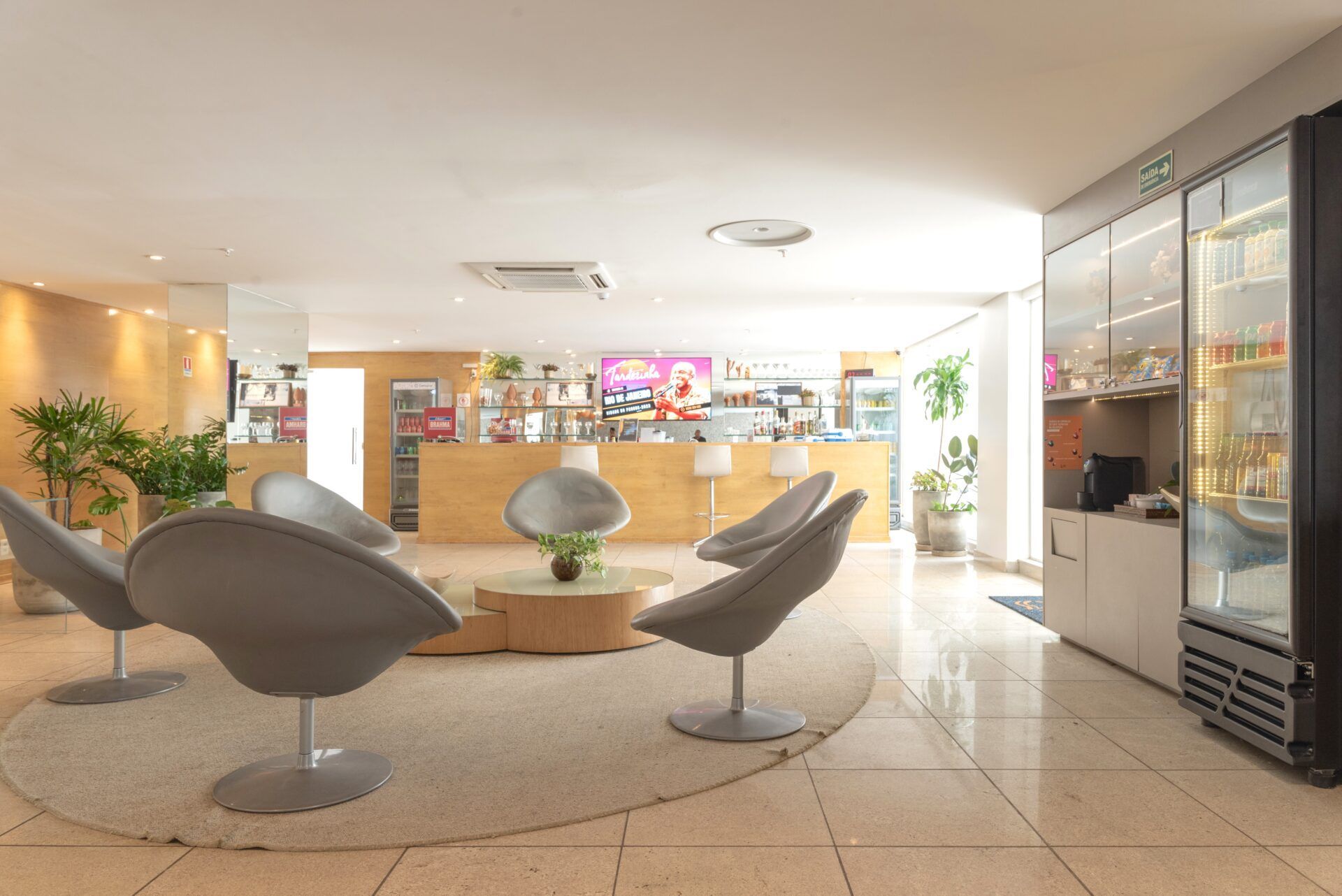 Lounge com sofás azul-petróleo e cadeiras rosa ao redor de uma mesa de madeira. Plantas suspensas e palmeiras altas em ambientes internos.