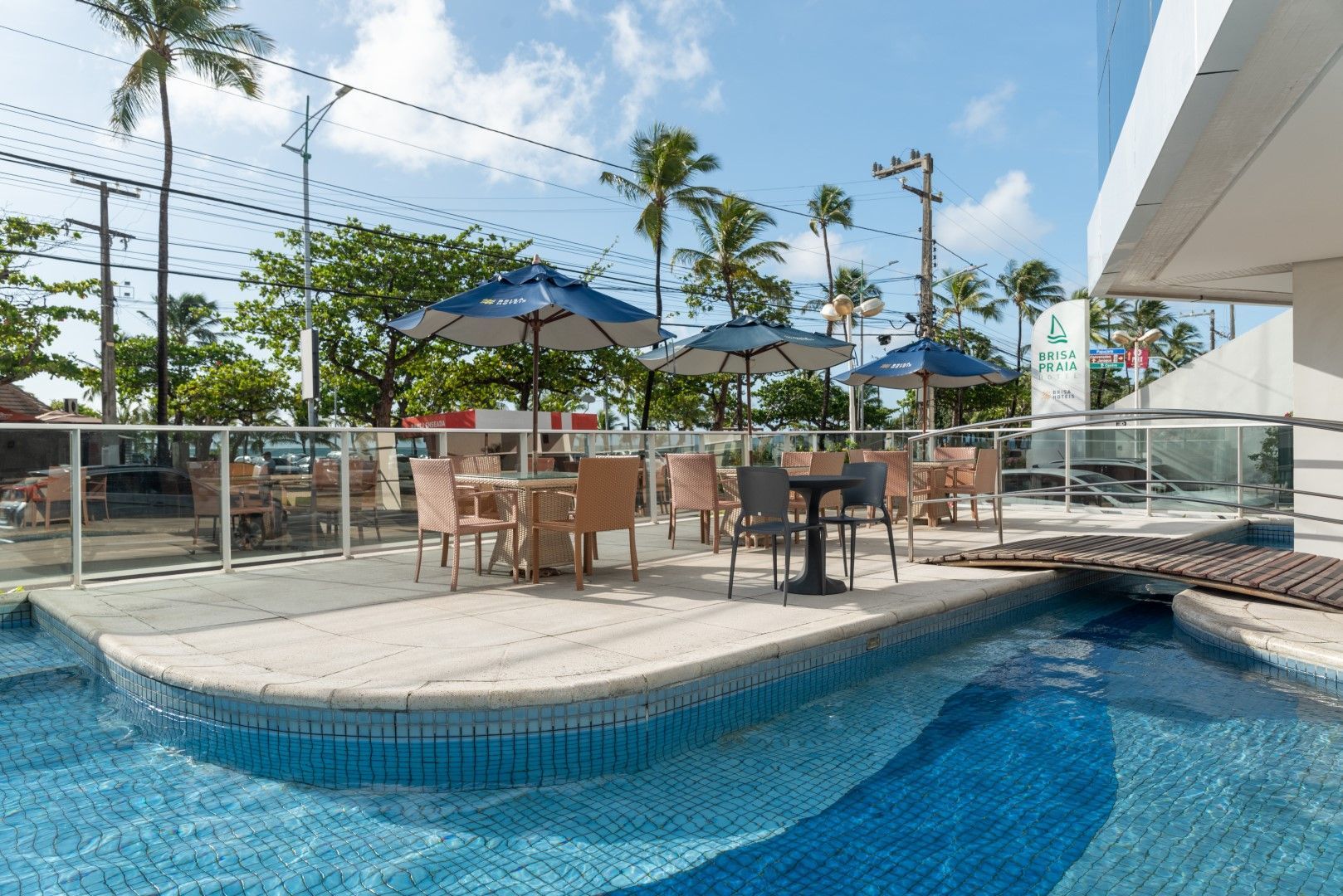 Café ao ar livre com mesas, cadeiras e guarda-sóis perto da piscina, com o oceano visível ao fundo.