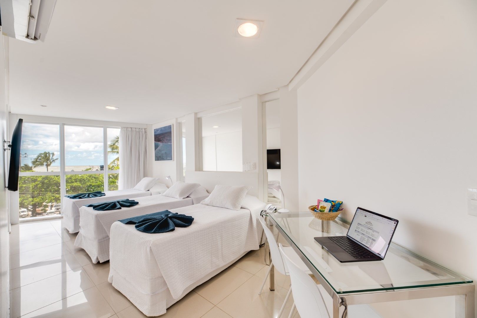 Quarto de hotel bem iluminado com duas camas, mesa com laptop e vista para o mar.