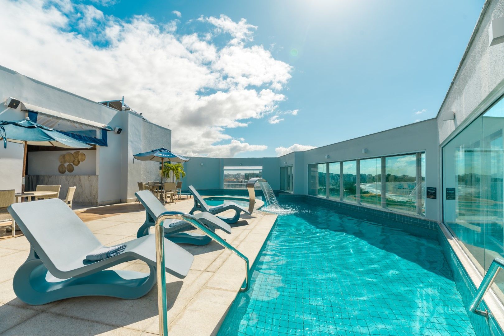 Deck de madeira com mesas, cadeiras e redes sob um guarda-sol perto da praia com palmeiras.