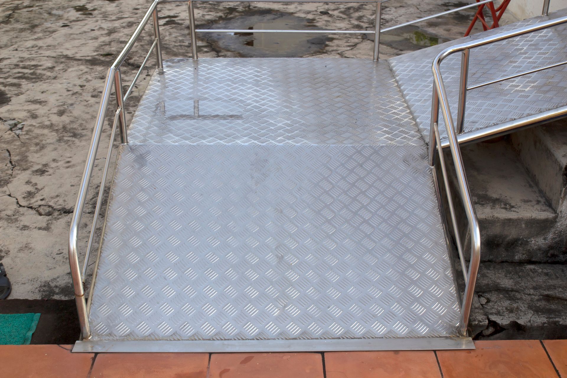 Una rampa metallica per sedie a rotelle con superficie a griglia diamantata e corrimano argentati, che conduce a una zona di atterraggio pavimentata.