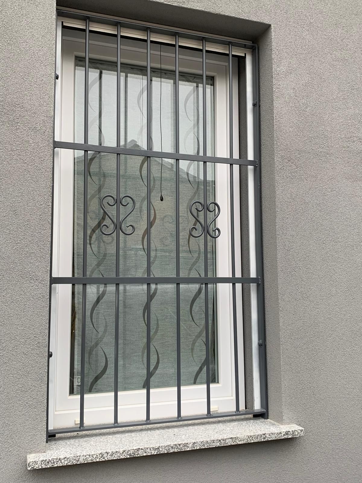 Una finestra con cornice bianca incastonata in una parete di stucco grigio, protetta da sbarre di sicurezza in ferro scuro con decorazioni a volute metalliche.