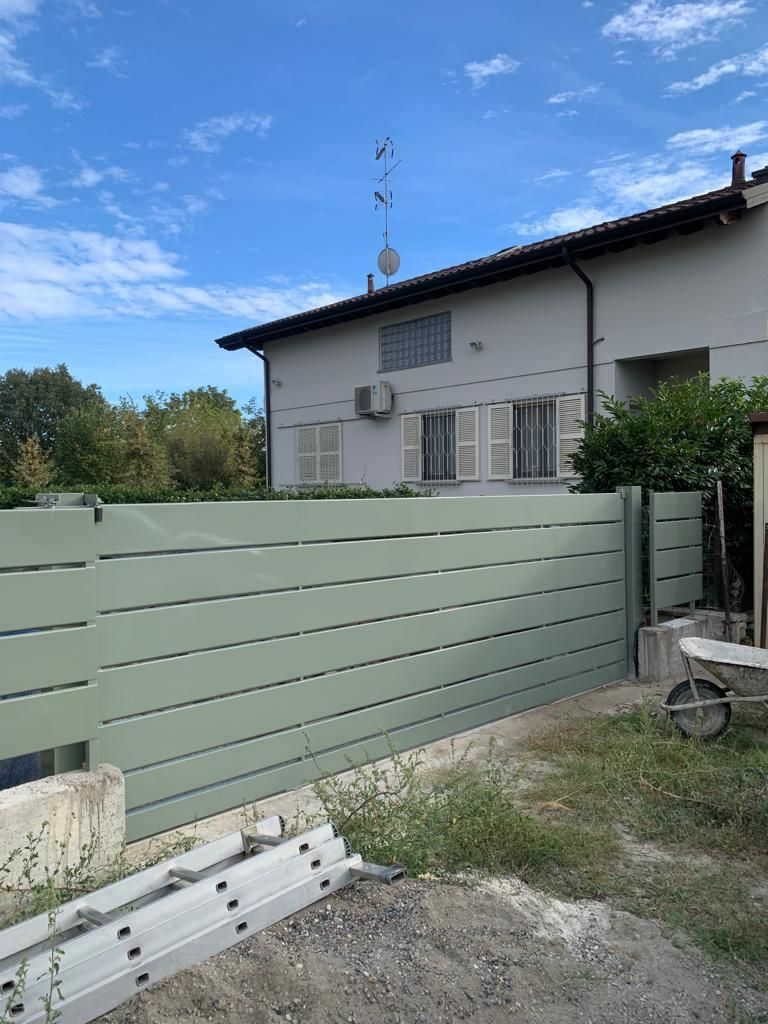 Una moderna recinzione orizzontale verde chiaro si erge di fronte a una casa bianca a due piani, sotto un cielo azzurro con nuvole sparse.
