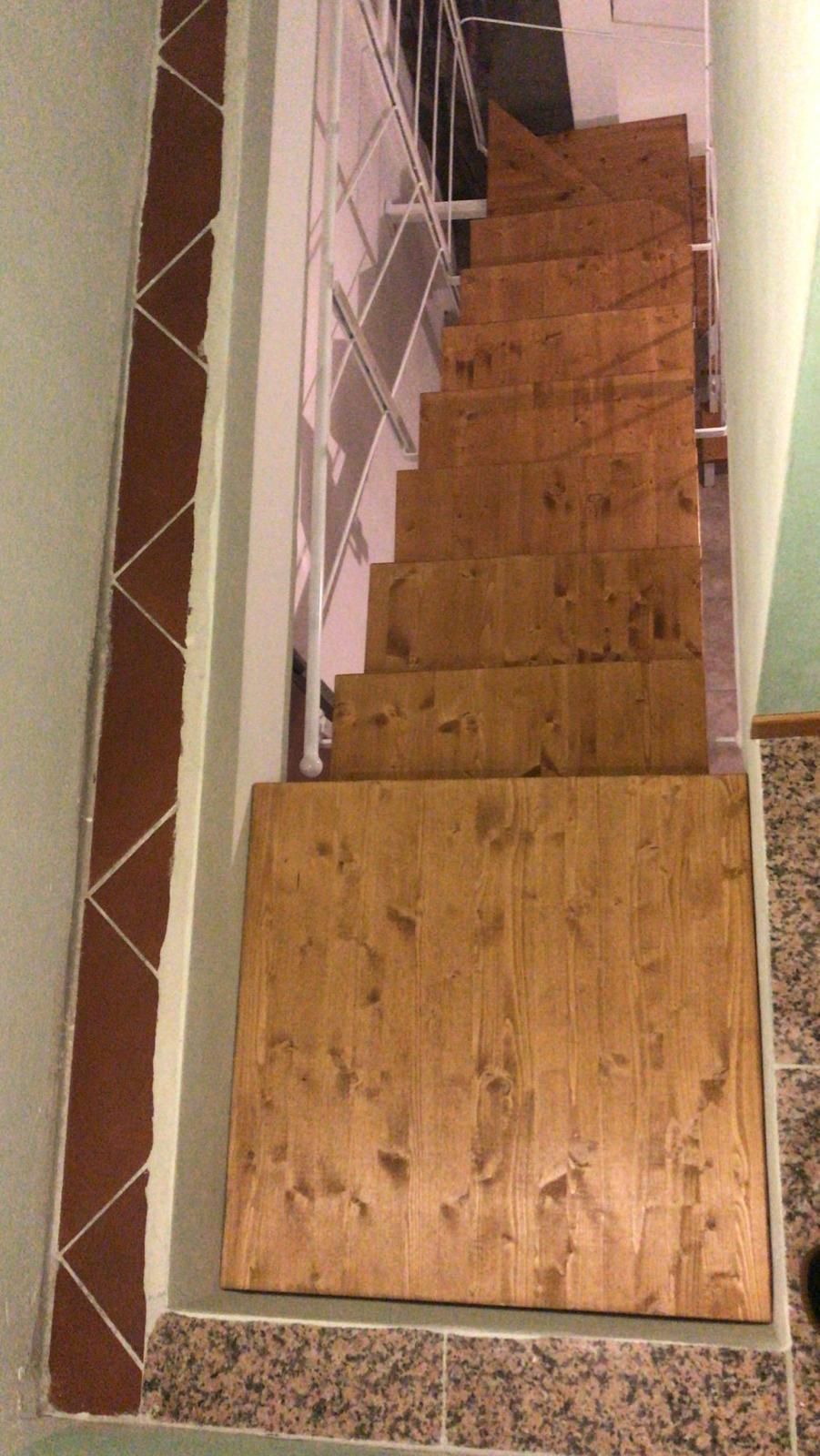 Una stretta scala con gradini in legno che scendono, fiancheggiata da una parete rivestita di piastrelle decorative marroni.