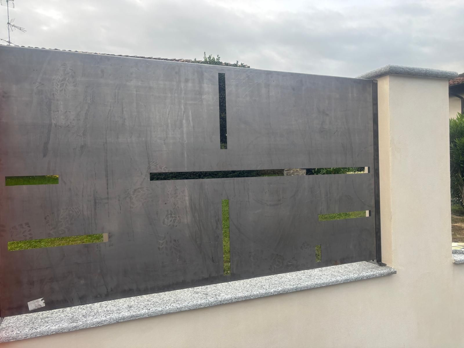 Un pannello di recinzione metallica con intagli orizzontali e verticali, sullo sfondo di vegetazione verde, montato su un muro di pietra.