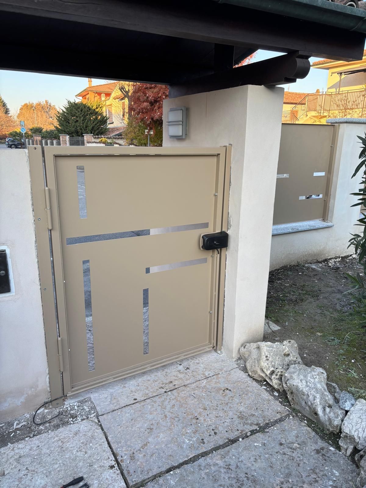 Un cancello pedonale in metallo beige con motivi geometrici argentati intagliati, incastonato in un muro di stucco in una zona residenziale esterna.