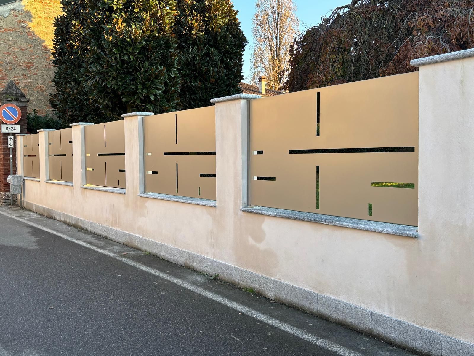 Vista a livello stradale di un muro in pietra beige con pannelli metallici moderni, equidistanti tra loro e caratterizzati da intagli geometrici.