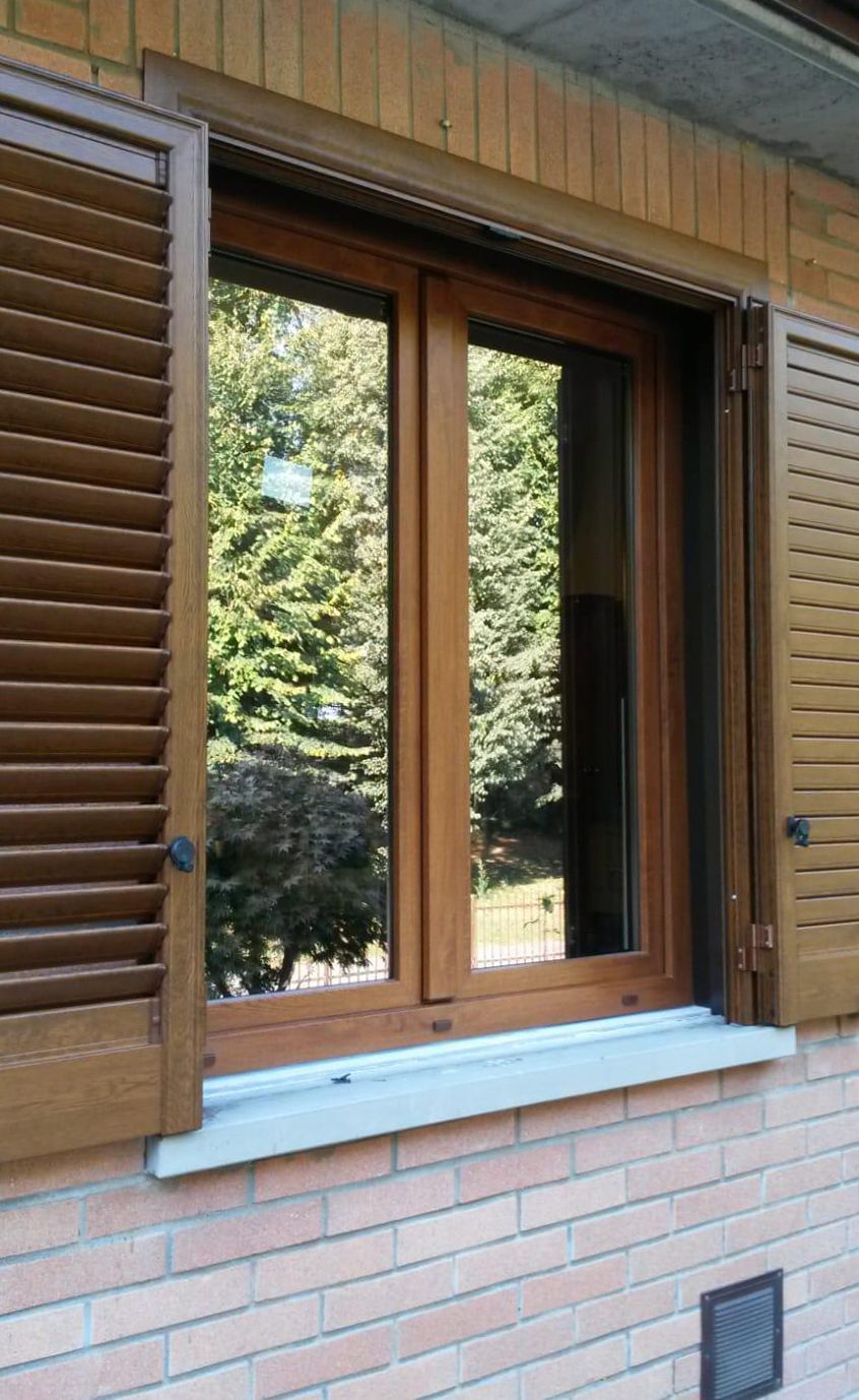 Una finestra in legno con persiane a lamelle aperte, incastonata in un muro di mattoni chiari, che si affaccia su una zona verde alberata.