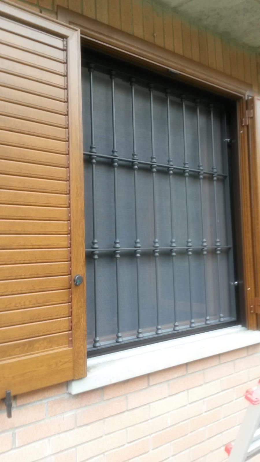 Una persiana di legno parzialmente aperta con lamelle orizzontali accanto a una finestra protetta da una grata di sicurezza in metallo scuro.