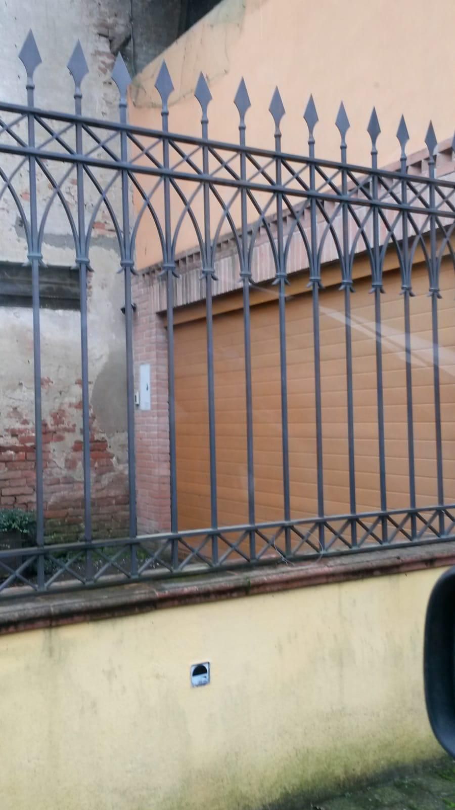 Una recinzione in ferro battuto nero con terminali a forma di diamante, posta sopra un muro giallo di fronte a una porta di garage e a un muro di mattoni.