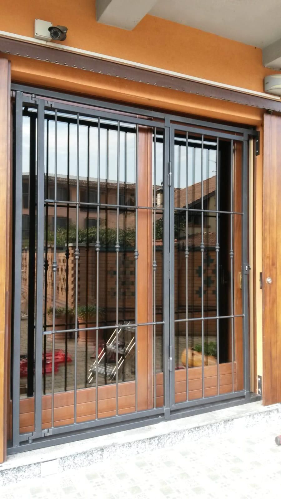 Un cancello di sicurezza in metallo scuro copre una serie di porte scorrevoli in legno e vetro su un patio con pavimento piastrellato.