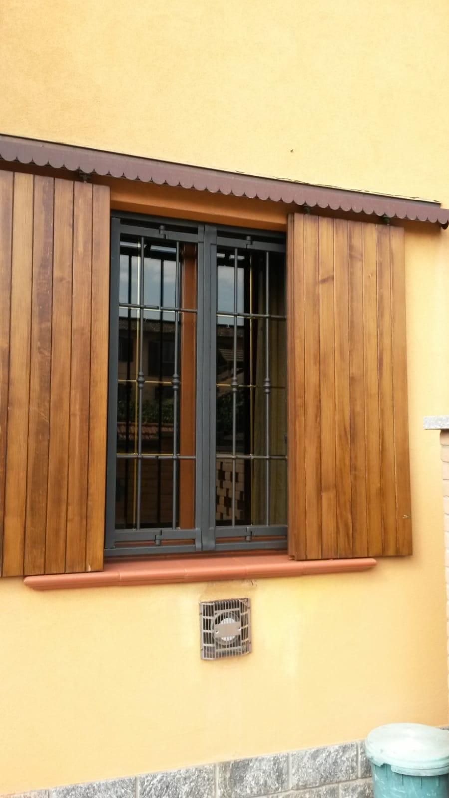 Una finestra con inferriate di sicurezza in metallo scuro e persiane color legno su una parete giallo pallido, con una piccola piastrella decorativa.