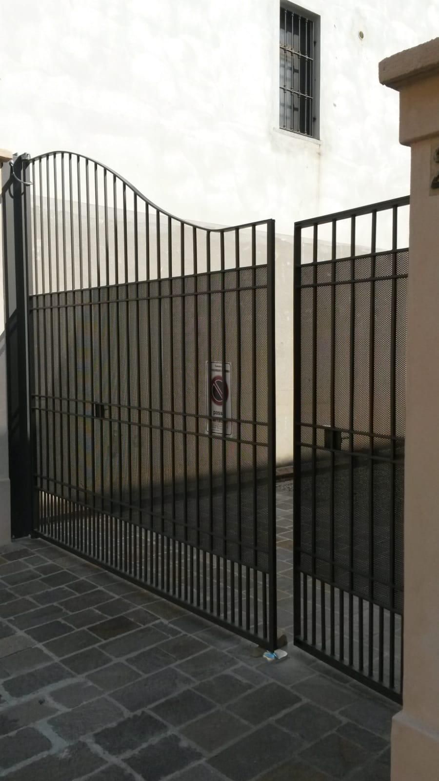 Un cancello doppio in metallo nero con la parte superiore curva, incastonato tra muri in pietra di fronte a un edificio bianco.