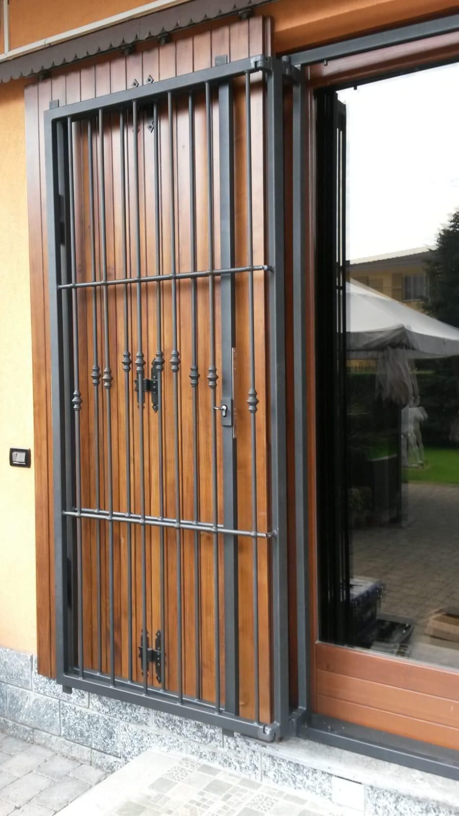 Un cancello di sicurezza in metallo scuro è socchiuso davanti a un telaio di porta in legno, lasciando intravedere un patio esterno.