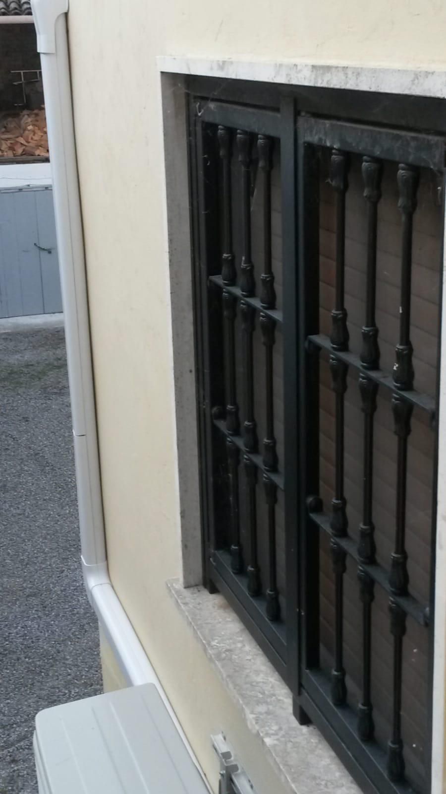 Una grata di sicurezza in metallo nero sopra una finestra sulla facciata di un edificio di colore chiaro, con un pluviale nelle vicinanze.