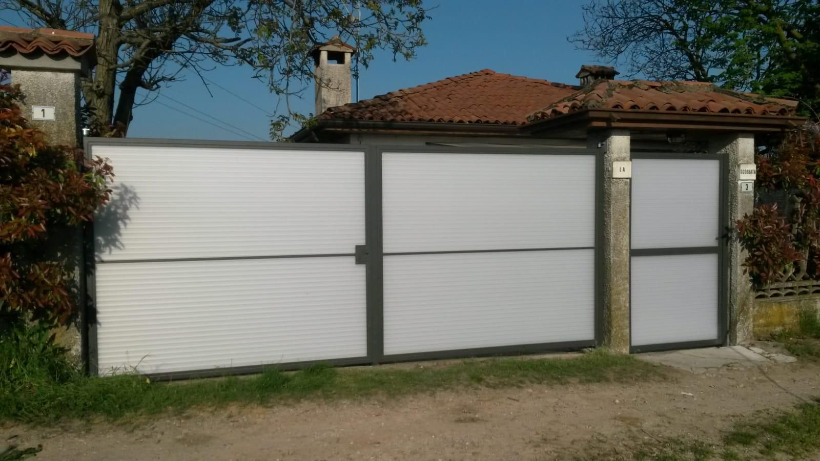 Un moderno cancello scorrevole a lamelle orizzontali color argento, con un cancello pedonale abbinato, incastonato tra pilastri in pietra in un contesto rurale.