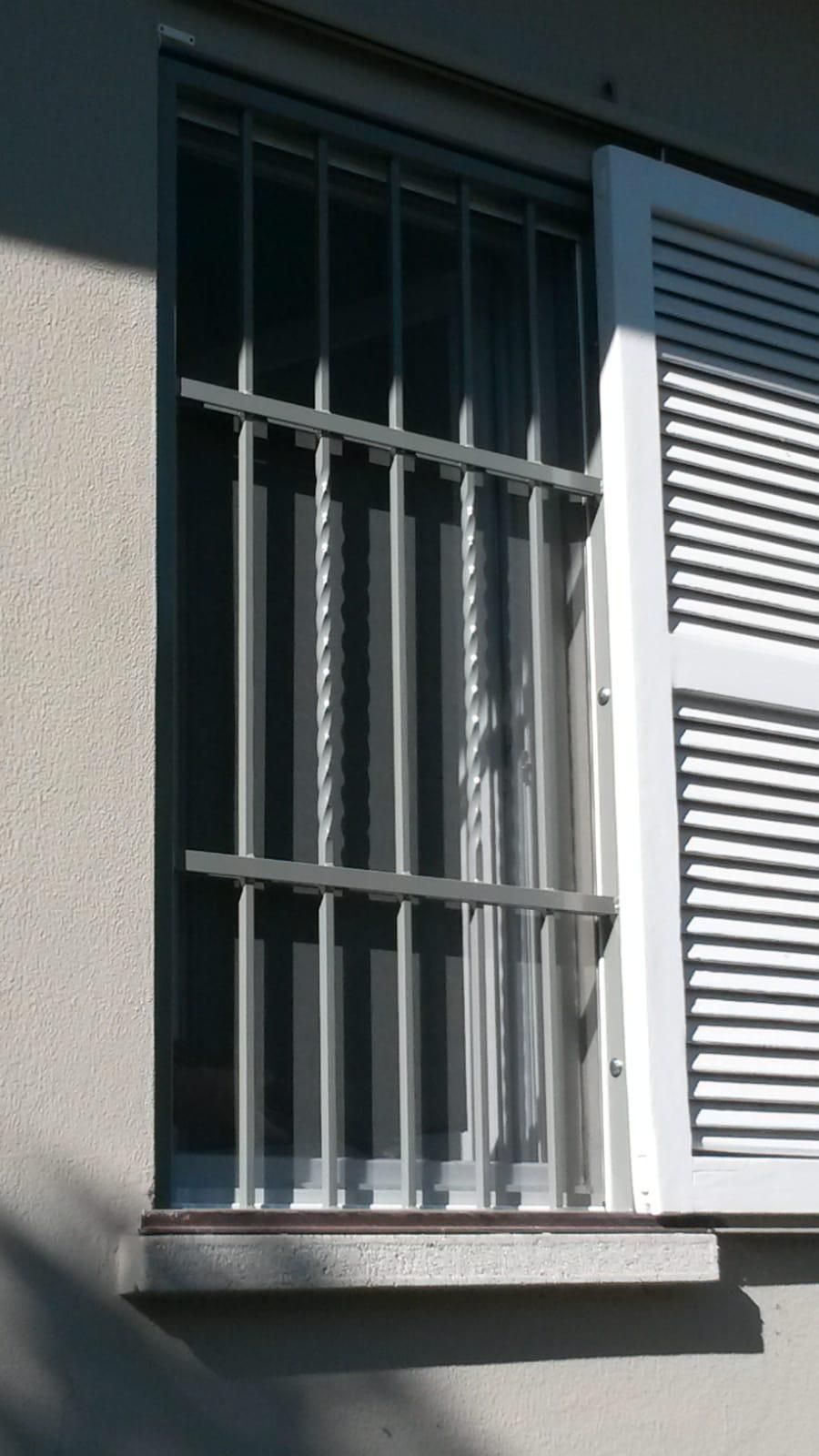 Una finestra grigia con sbarre di sicurezza verticali e una persiana a lamelle bianche sul lato, vista su uno sfondo grigio chiaro.