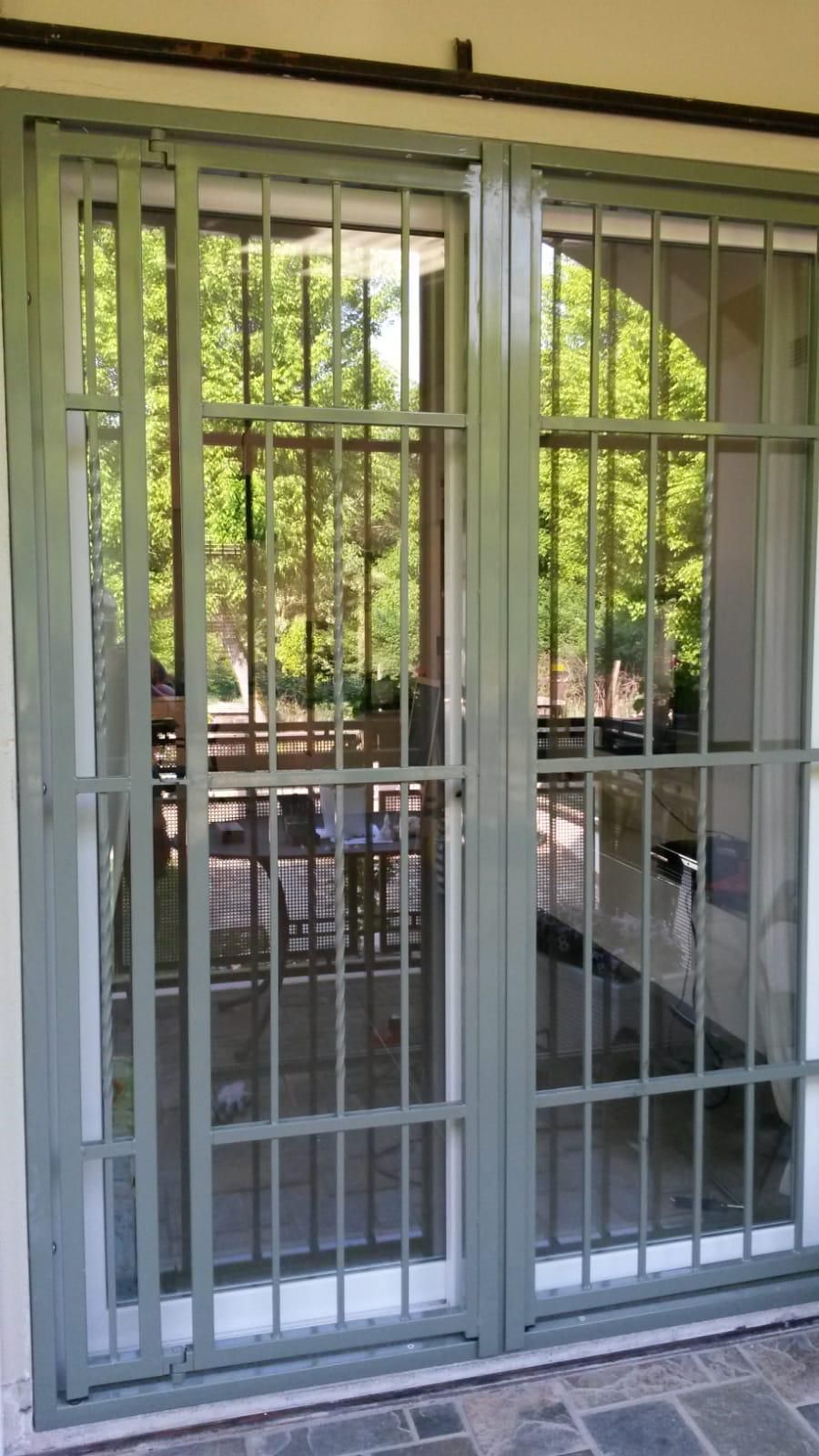 Un cancello di sicurezza in metallo grigio con motivo a griglia, installato sopra una porta a vetri che lascia intravedere un patio esterno sfocato.