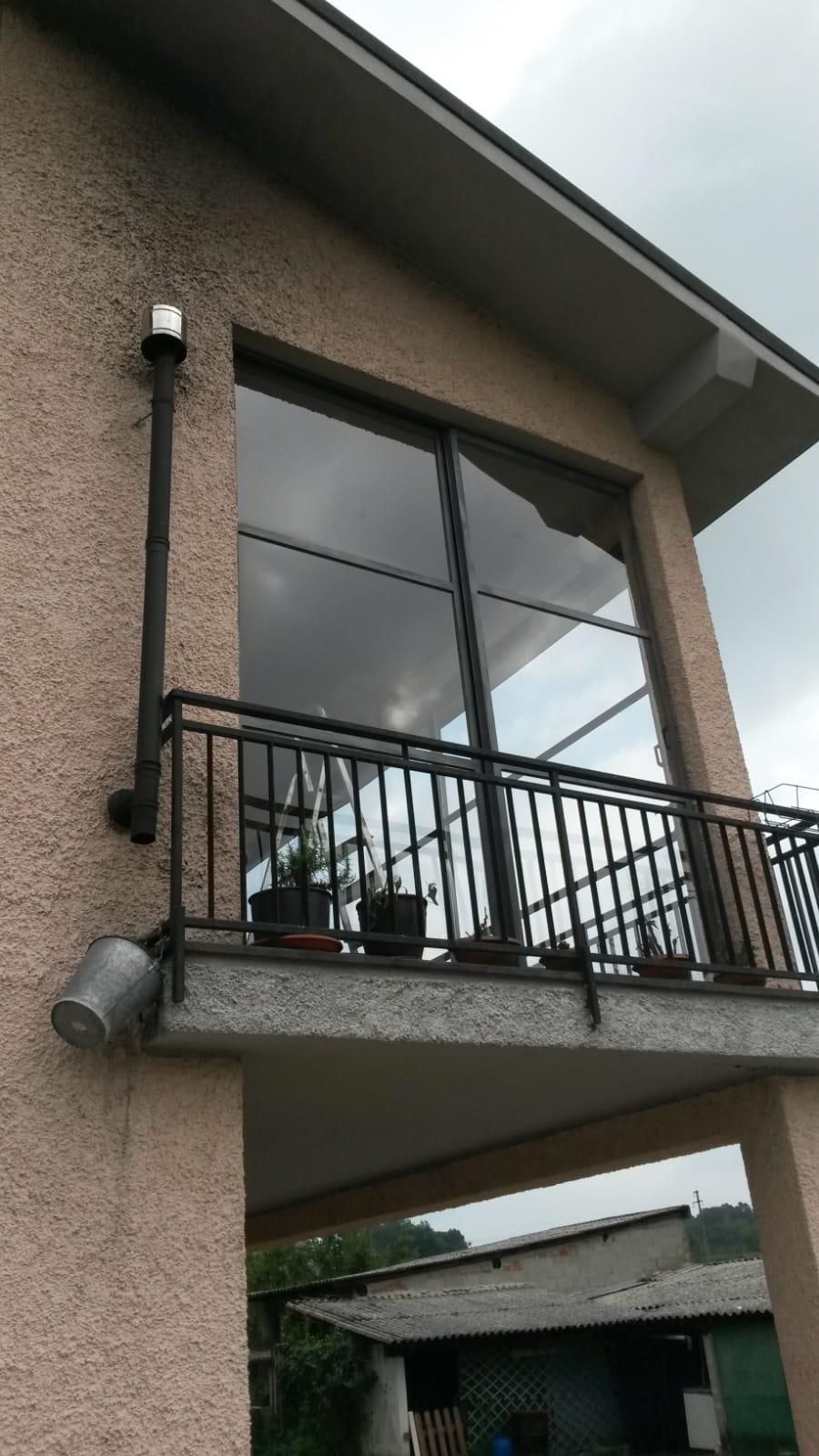 Esterno di una casa beige con un piccolo balcone, caratterizzato da un tubo di metallo scuro, una finestra e delle piante.