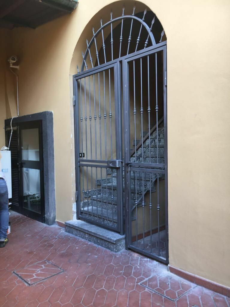 Un arco con un cancello di sicurezza in metallo scuro che conduce a una scala interna, sullo sfondo di pareti color beige chiaro.