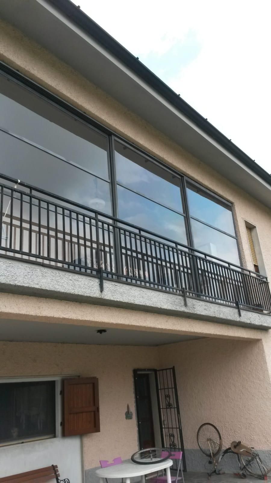 Esterno di un edificio a due piani con un balcone chiuso da ampie vetrate sopra un'area patio.