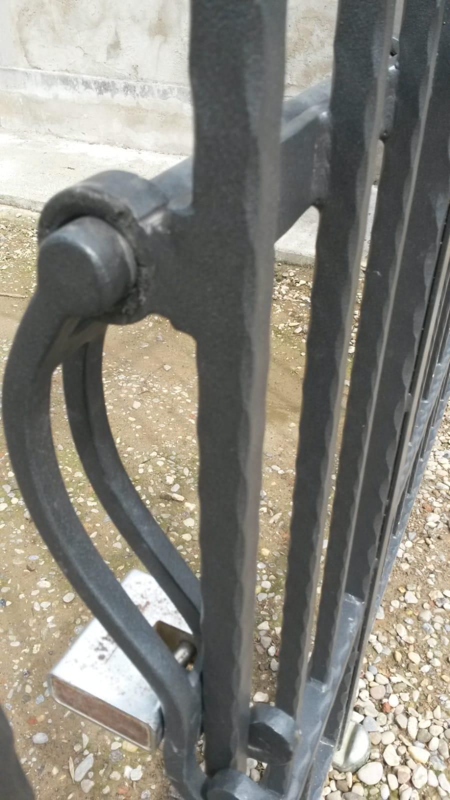 Primo piano di un cancello in ferro nero con superficie ruvida, maniglia curva e un pesante lucchetto in metallo chiuso nella parte inferiore.